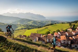 Vététiste et randonneur sur un chemin au-dessus d’un village basque, avec les collines et l’océan en arrière-plan